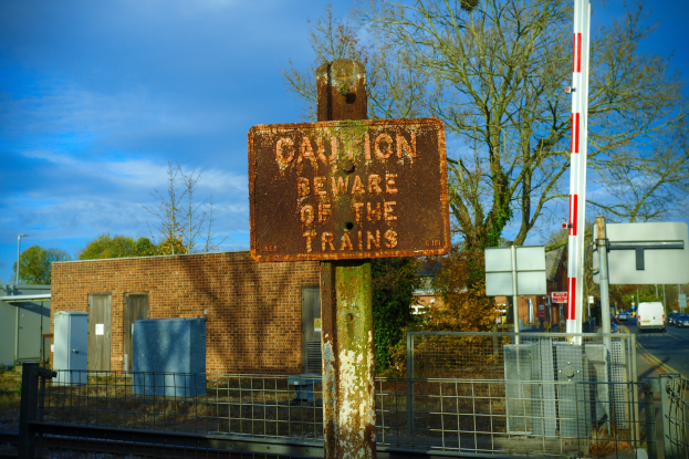 Vorsichtsschild an einer Bahnschiene umgeben von einem Metallzaun, Bäumen, Strommasten, einem Gebäude mit Fenstern, Containern, Fahrzeugen auf einer Straße und einem bewölkten Himmel.