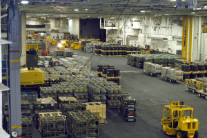 Lagerhalle mit mehreren Lastwagen, Containern, Menschen, Deckenleuchten, Metallstangen und einem sichtbaren Rad.