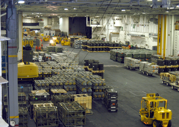 Lagerhalle mit mehreren Lastwagen, Containern, Menschen, Deckenleuchten, Metallstangen und einem sichtbaren Rad.