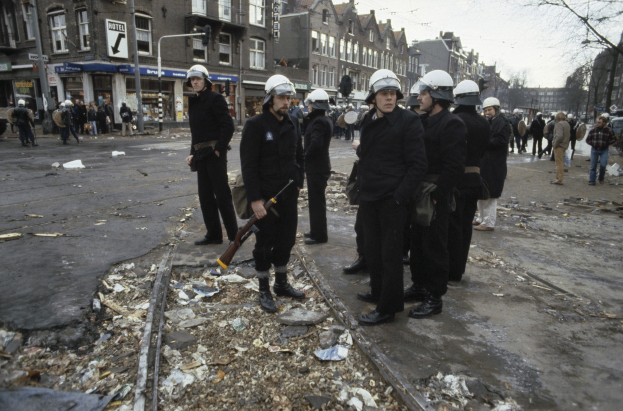 Eine Gruppe von Polizisten in Helmen und mit Waffen in der Hand steht an der Straße, mit Gebäuden, Schildern, Laternen, Bäumen und einem klaren Himmel im Hintergrund.