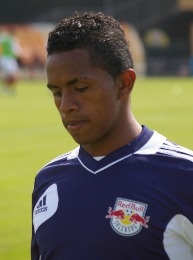 Junger Mann in blauem T-Shirt auf einem Fußballfeld, der wie ein Flügelstürmer mit kurzen dunklen Haaren und entschlossener Miene erscheint.